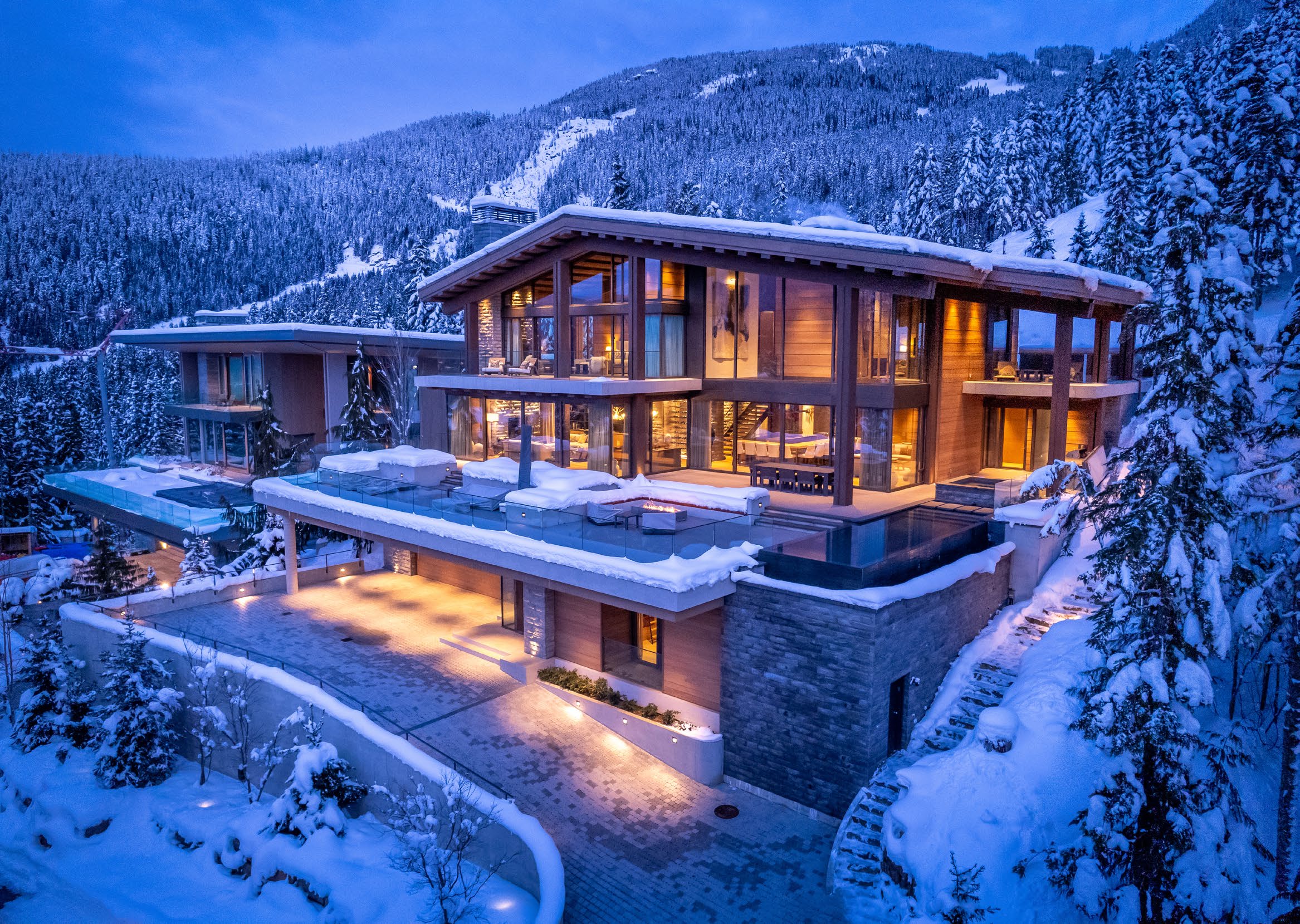 Illuminated timber and glass ski-in chalet with infinity pool in deep snow at dusk in Whistler, British Columbia